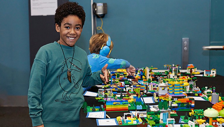 Child pointing at LEGO creations with another child in the background wearing headphones