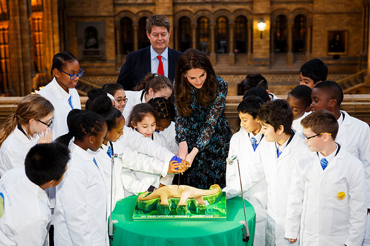 Photo of HRH The Duchess of Cambridge cutting a Diplodocus-shaped cake in the Museum with help from some schoolchildren